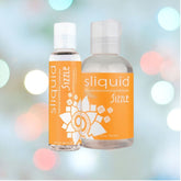 Two bottles of Sliquid Naturals 'Sizzle' Warming Lubricant are showcased against a gently blurred, vibrant backdrop. The packaging displays an orange label adorned with white text and intricate designs, emphasizing its water-based formula with a hint of menthol.